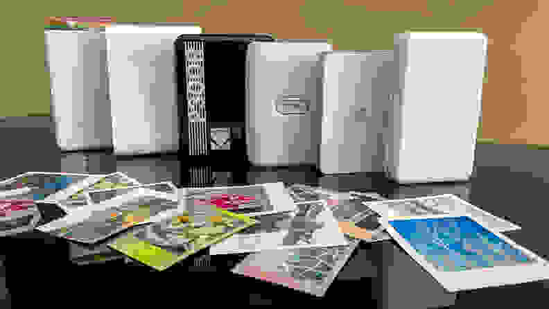 The Polaroid Hi-Print, Canon Ivy, Kodak Step, Fujifilm Instax Mini Link, Lifeprint 2x3 Hyperphoto, and Victure PT320 portable photo printers lined up on a table with a collection of photos in front of them.