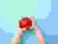 A toddlers hands extended holding a red apple against a light blue background.