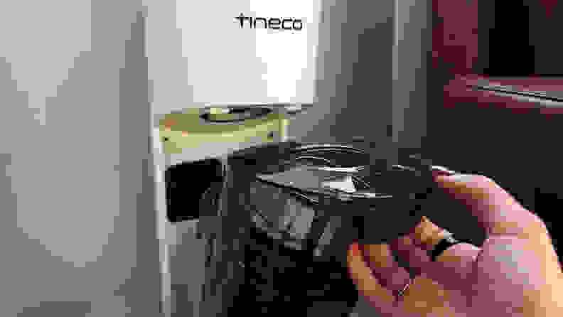A hand removes a waste bin from a white Tineco vacuum base.