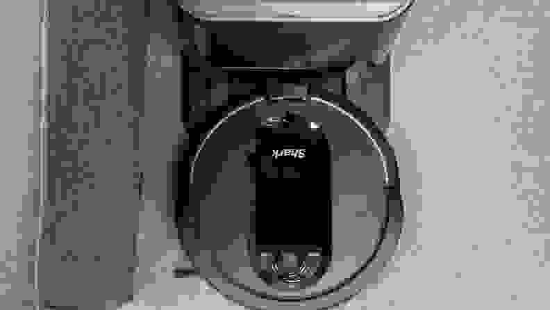 A circular black Shark IQ robot vacuum trundles along on the carpet. We're looking down at it from above.