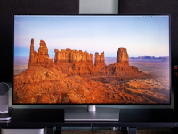 A widescreen monitor displays a high-resolution image of red rock formations in a desert landscape, framed by black studio speakers on a sleek desk setup.