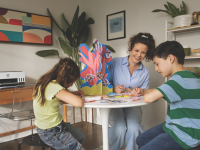 A woman and two children smile and work on a colorful arts and crafts project at a round table in a cozy, plant-filled home office with an HP printer in the background.